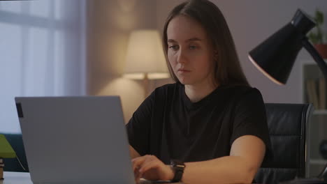 young-female-writer-or-journalist-is-typing-text-on-laptop-working-in-home-at-evening-sitting-at-table-in-room-of-apartment-portrait-of-freelancer-woman-at-work